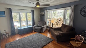 Un salon avec un ventilateur de plafond, deux fenêtres avec des rideaux, un canapé, un fauteuil, un tapis et un fauteuil à bascule en bois.