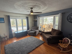 Un salon avec un ventilateur de plafond, deux fenêtres avec des rideaux, un canapé, un fauteuil, un tapis et un fauteuil à bascule en bois.