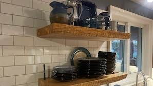 Une cuisine aux murs recouverts de carreaux blancs, avec deux étagères en bois où sont posées des assiettes et des bols, et une fenêtre donnant sur des arbres.