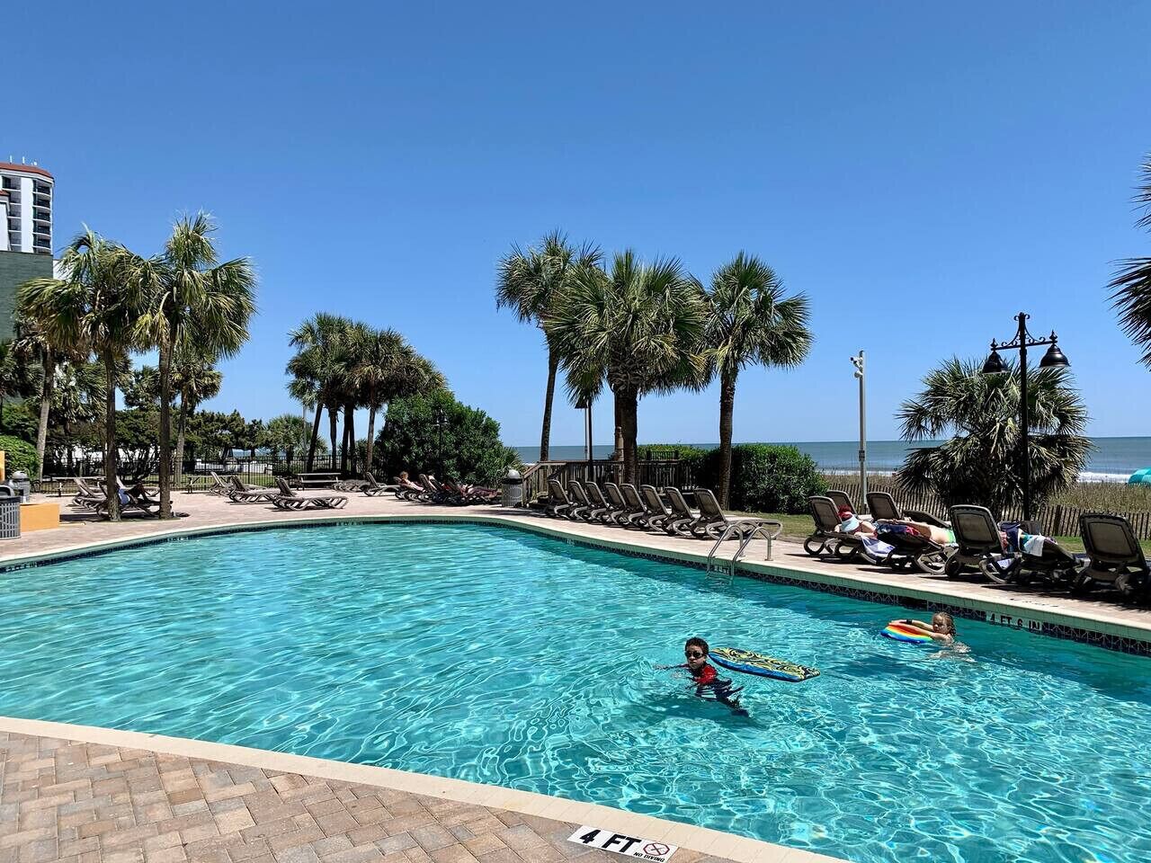 A swimming pool with lounge chairs and palm trees, a lifeguard tower, and a view of the ocean.
