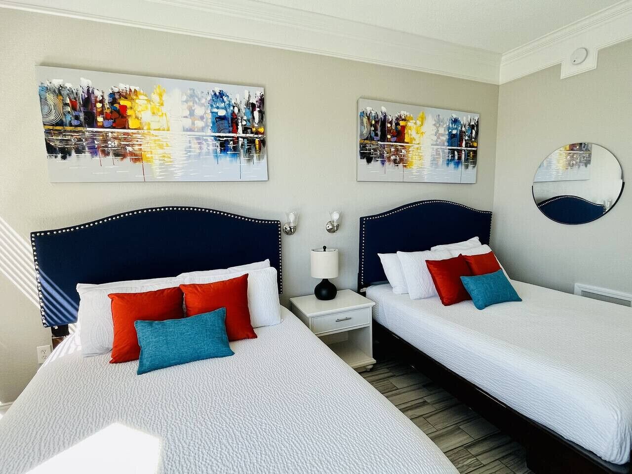 Une chambre d’hôtel avec deux lits, chacun doté d’une tête de lit, de draps blancs et d’oreillers rouges et bleus. Deux œuvres d’art sont accrochées au mur et représentent des paysages urbains, et un miroir rond est également fixé au mur.