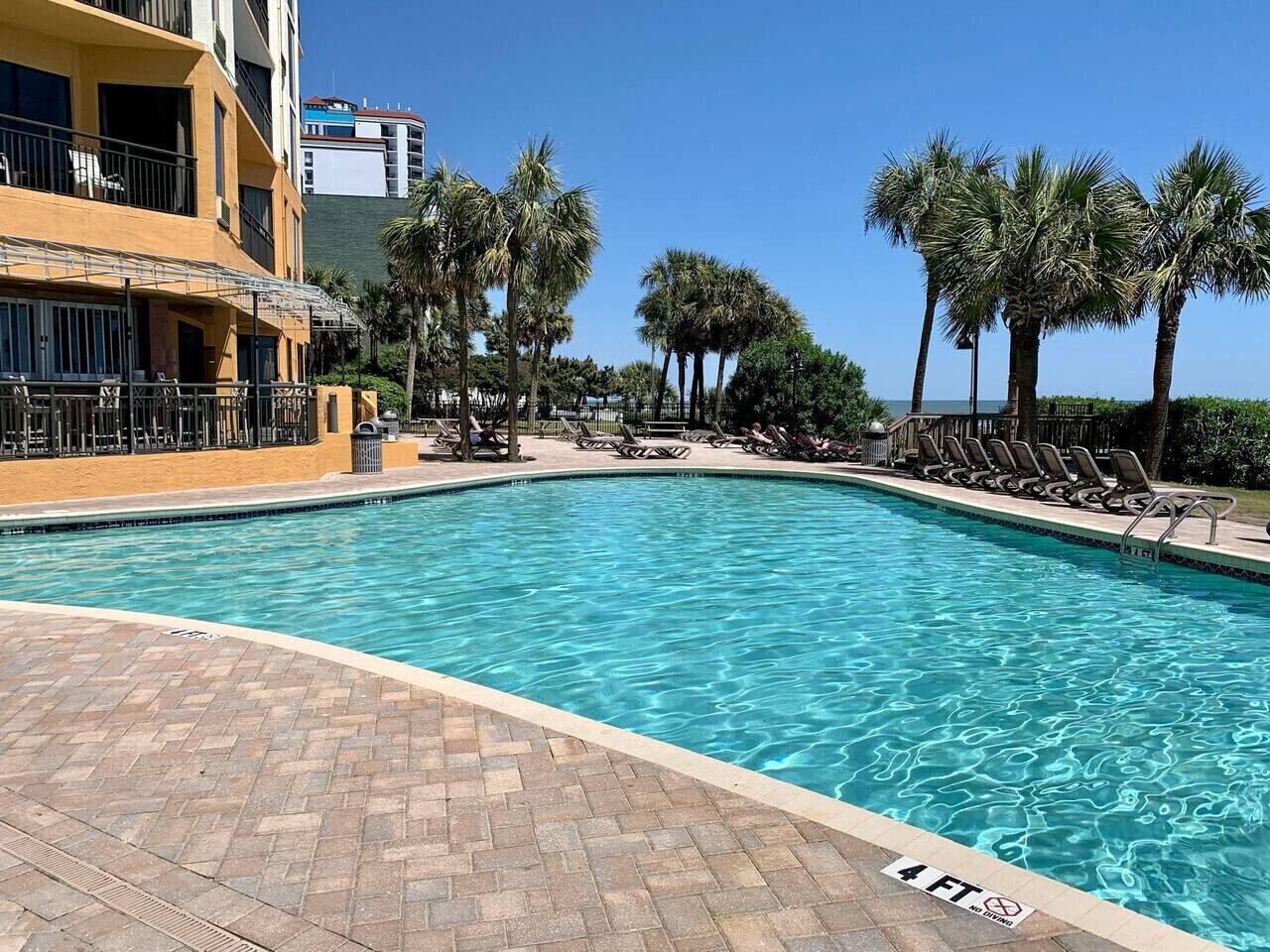 A swimming pool with lounge chairs and palm trees, surrounded by a brick patio and a building with balconies.