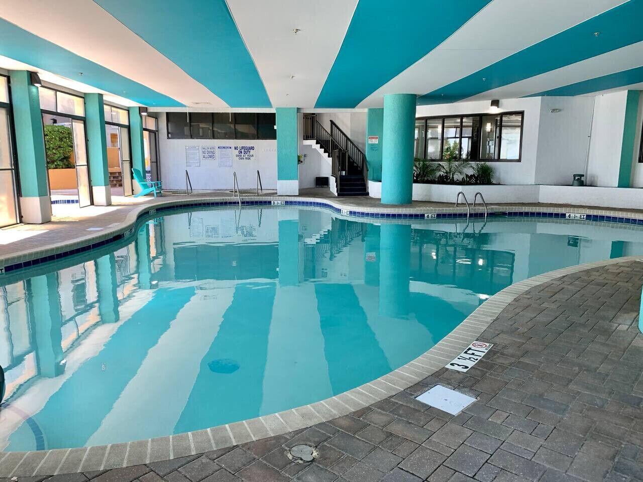 An indoor swimming pool with clear blue water, a tiled deck, and a staircase leading to an upper level.