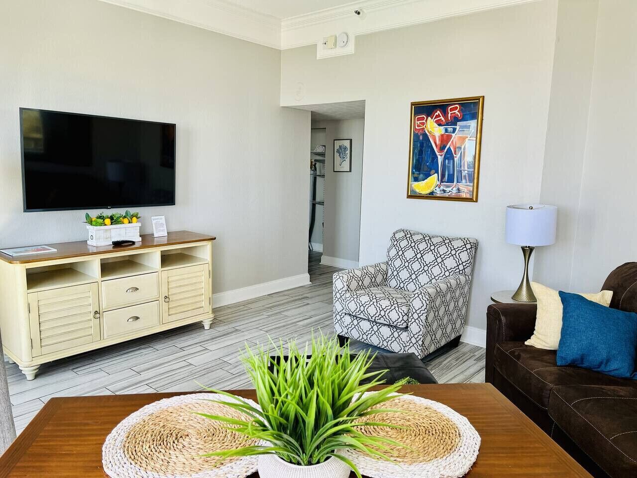 A living room with a flat-screen TV, a patterned armchair, a brown sofa, and a coffee table with a plant.