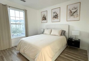 Een slaapkamer met een groot bed, een nachtkastje, een raam met gordijnen en drie ingelijste kunstwerken aan de muur.