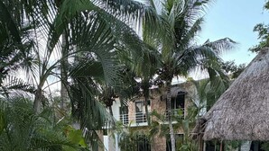 A pool area with a thatched-roof structure, palm trees, and a stone building in the background. - Luxury Studio & 1BR Condos • Pools • Near Tulum & Akumal Beaches (Akumal)
