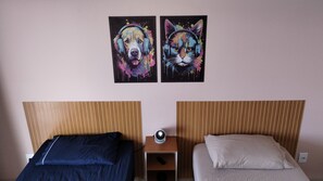 Una habitación con dos camas, cada una con cabecera, una mesita con un altavoz, y dos cuadros colgados en la pared de un perro y un gato.