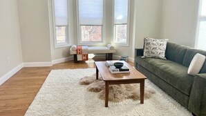 A living room with a wooden coffee table, a green sofa, a white shag rug, and a white bench against a wall with three windows. - Broadway Nomad House in Providence (Providence)