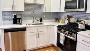 A modern kitchen with white cabinets, a stainless steel dishwasher, a coffee maker, a kettle, and a microwave. - Broadway Nomad House in Providence (Providence)