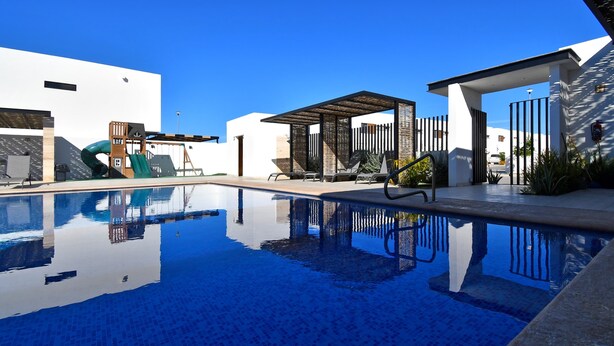 Uma piscina com superfície de água azul cristalina, rodeada por um ambiente arquitetônico moderno, com área de estar coberta e um parquinho infantil.