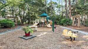Un terrain de jeux avec des toboggans et des balançoires, entouré d’arbres et agrémenté d’un espace jardin avec des plantes en pot.