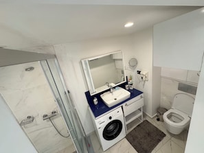 A modern bathroom with a glass-enclosed shower, a sink with a mirror above it, a washing machine, and a toilet. - Orbi City Apartaments  (Batumi)