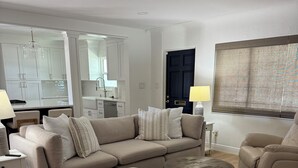 Een moderne woonkamer met een hoekbank, een poef en een ronde salontafel. Achtergrond toont een keuken met witte kastjes en een zwarte deur.