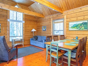 L’intérieur chaleureux d’un chalet avec une table à manger en bois, des chaises, un canapé et une grande fenêtre donnant sur la forêt.