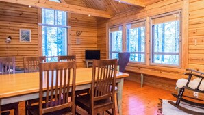 A wooden cabin interior with a dining table, chairs, a television, and large windows offering a view of the forest. - Luhtalemmikki by Interhome (Lieksa)