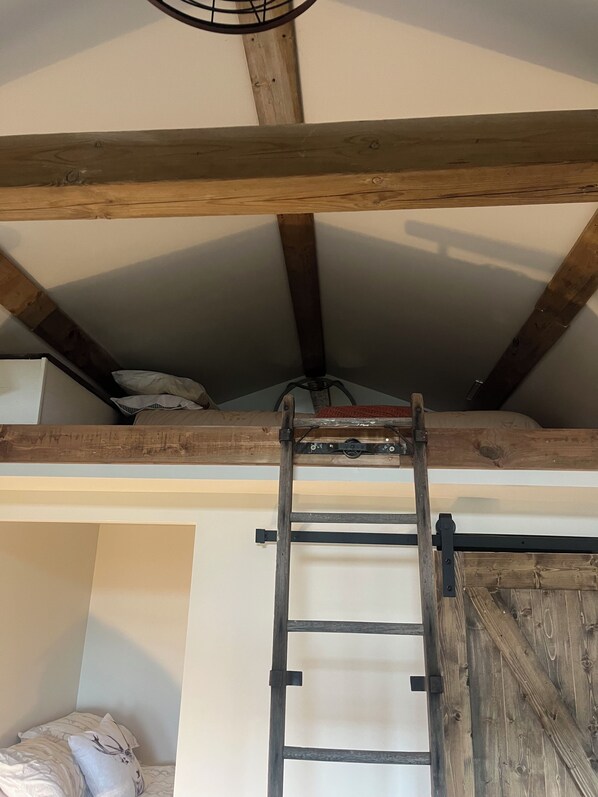 A loft-style bedroom with exposed wooden beams, a bunk bed, a ladder, and a small window. - Wake up to the beautiful sunrise on Lake Erie in this tiny lakeside cabin. (Wheatley)