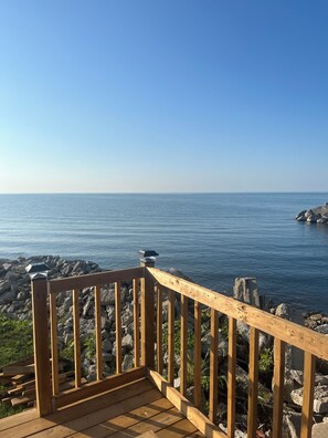 A wooden deck with a railing overlooking a calm sea and rocky shoreline. - Wake up to the beautiful sunrise on Lake Erie in this tiny lakeside cabin. (Wheatley)