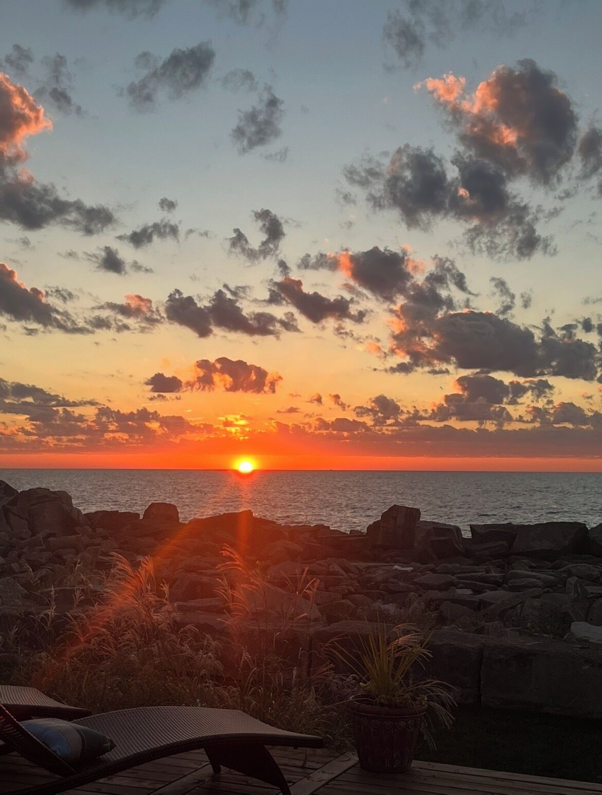 A sunset over a rocky coastline with a lounge area in the foreground.