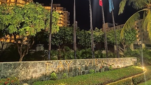 Scena notturna con un muro di pietra su cui è inciso "BAY VIEW GRILL" e diverse bandiere, tra cui quella americana, di fronte a un edificio con finestre illuminate.