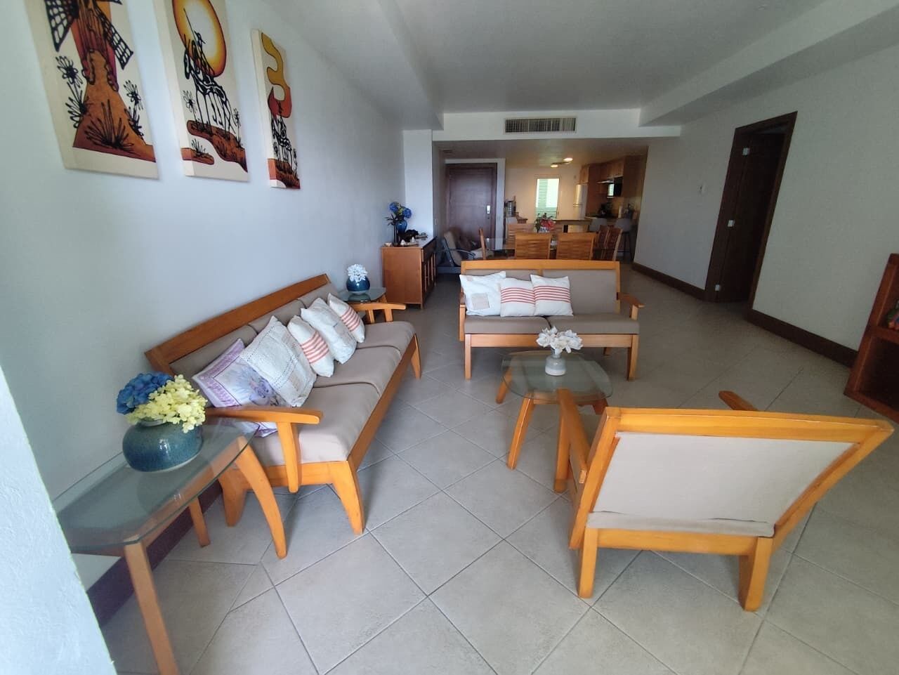 A spacious living area with a sofa set, a coffee table, and a dining table. There are wall-mounted artworks and a door leading to another room.