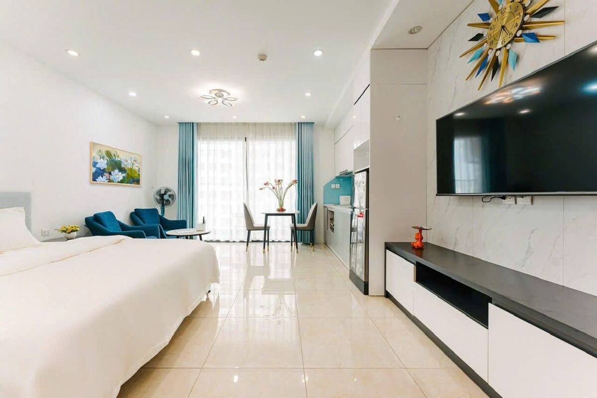 A modern hotel room with a large bed, a dining area with a table and chairs, a kitchen area with a refrigerator, and a mounted TV.