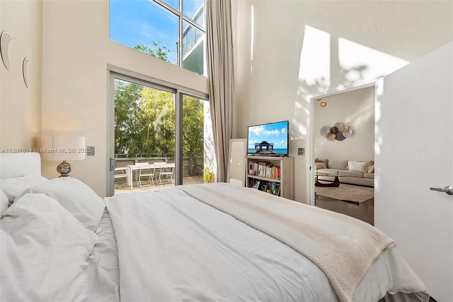 A bedroom with a bed, a TV, a bookshelf, and a balcony with a view of trees.