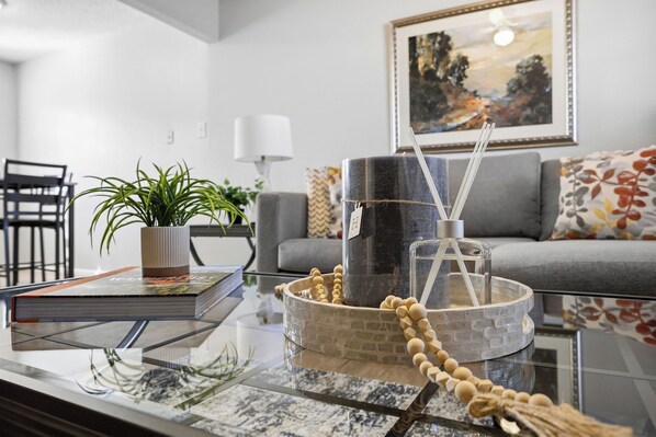 A modern living room with a glass coffee table, a potted plant, a book, a decorative bowl, and a framed painting on the wall.
