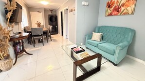 Ein Wohnzimmer mit einem petrolfarbenen Sofa, einem Glastisch, einem Essbereich mit Stühlen und einem an der Wand montierten Fernseher.