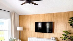 A modern living room with a ceiling fan, a flat-screen TV mounted on a wooden wall, a white cabinet, and a patterned rug. - North Boulevard Breeze Stylish New Home Near Beaches & Historic District (St. Augustine)