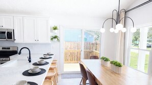 A modern kitchen with white cabinets, a central island with stools, and a dining area with a long wooden table and chairs. - North Boulevard Breeze Stylish New Home Near Beaches & Historic District (St. Augustine)