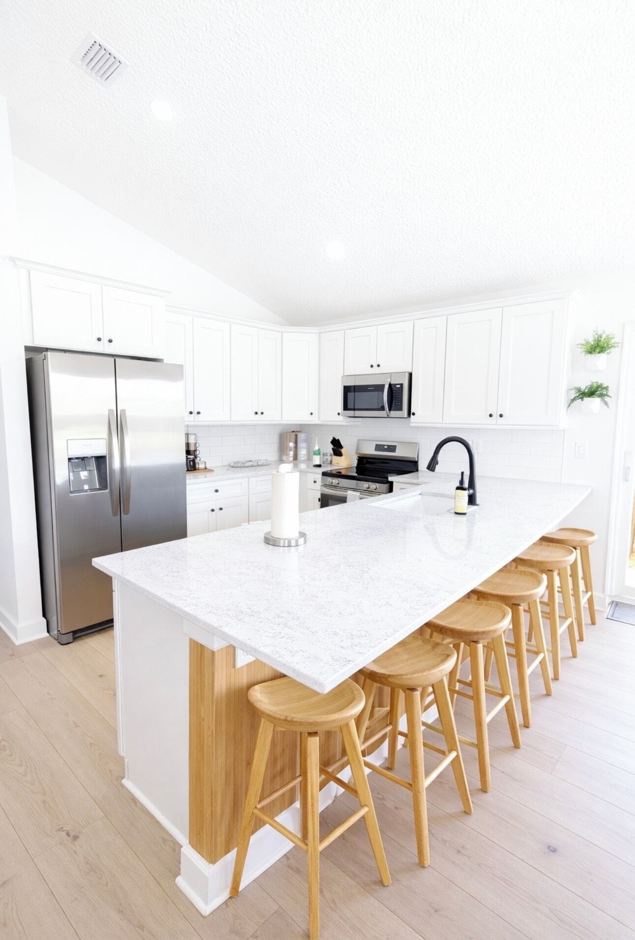 A modern kitchen with white cabinetry, stainless steel appliances, a large island with stools, and wooden flooring.