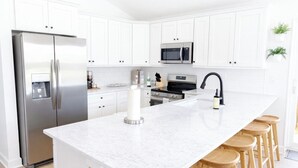 A modern kitchen with white cabinetry, stainless steel appliances, a large island with stools, and wooden flooring. - North Boulevard Breeze Stylish New Home Near Beaches & Historic District (St. Augustine)
