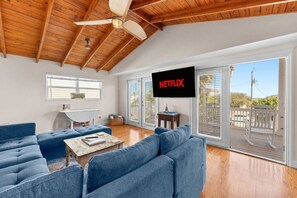 Ein geräumiges Wohnzimmer mit einem großen Fernseher, der 'NETFLIX' zeigt, einer Holzbalkendecke und einem Balkon mit einem weißen Stuhl.