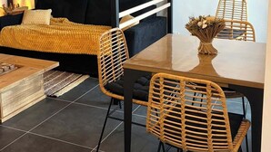 Una cama litera con estructura negra y barandillas blancas, un sofá negro con una manta amarilla, dos sillas de mimbre, una mesa de centro de madera y un jarrón con flores sobre un piso de baldosas.