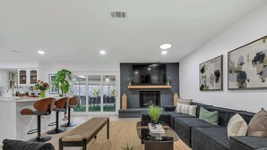A modern living room with a black sofa, a glass coffee table, and a fireplace. There are three wooden chairs and a bench. The room has a large window and a kitchen area in the background. - Indoor Pool & Hot Tub Near AT&T Stadium (Irving)