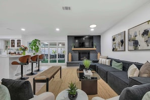 A modern living room with a black sofa, a glass coffee table, and a fireplace. There are three wooden chairs and a bench. The room has a large window and a kitchen area in the background. - Indoor Pool & Hot Tub Near AT&T Stadium (Irving)