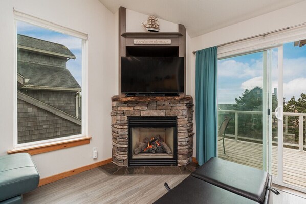 A modern living room with a stone fireplace, a flat-screen TV mounted above it, and a large sliding glass door leading to a balcony. - Pelican Watch - - Block from Beach w/Hot Tub Dog Friendly (Cloverdale)