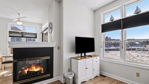 A living room with a fireplace, a television on a cabinet, and a view of a snowy landscape through large windows. - Marina Studio- King Bed, No Steps (Manistee)