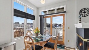 A dining area with a glass table, wooden chairs, a potted plant, and a view of a snowy landscape through large windows. - Marina Studio- King Bed, No Steps (Manistee)