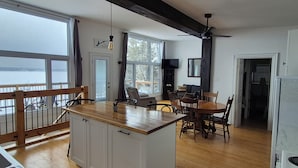Une cuisine avec des armoires en bois, un coin repas avec des chaises, un ventilateur de plafond et une grande fenêtre donnant sur une étendue d’eau.