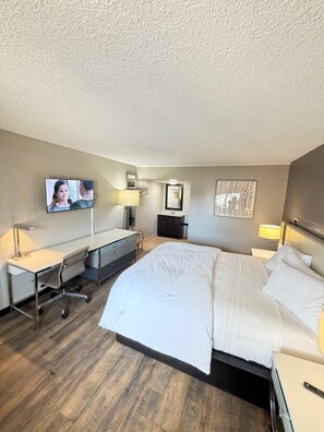 A hotel room with a bed, desk, chair, TV, and a wall with a framed picture. - Deluxe Inn (Mauldin)