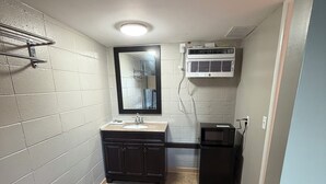 A small, well-lit bathroom with a sink, mirror, and vanity. There is a towel rack on the wall and an air conditioning unit mounted above the mirror. The floor is tiled with a patterned design. - Deluxe Inn (Mauldin)