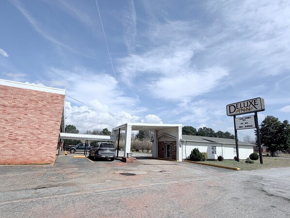 A hotel entrance with a sign for 'Deluxe Inn' and a parking lot with a few cars. - Deluxe Inn (Mauldin)