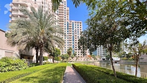 A pathway lined with neatly trimmed hedges and palm trees, leading to a body of water with a high-rise building in the background. - Vibrant 2 Bed l The Greens l Family Retreat (The Greens)