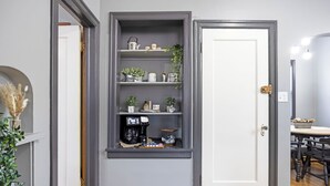 Una habitación con una puerta gris, una estantería con plantas y utensilios de cocina, y un comedor con una mesa y sillas.