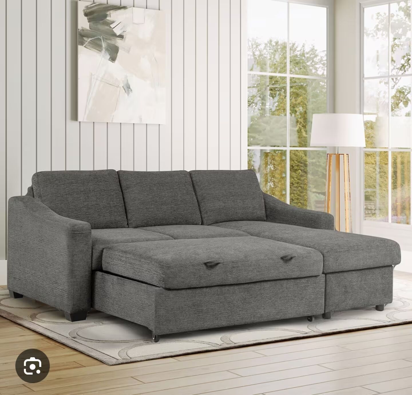 Un canapé d’angle gris avec une méridienne, placé sur un tapis de couleur claire dans une pièce aux murs recouverts de panneaux blancs et dotée de grandes fenêtres.