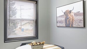 Un comedor con una mesa redonda, sillas negras, un centro de mesa decorativo, una ventana con persianas y un cuadro en la pared.