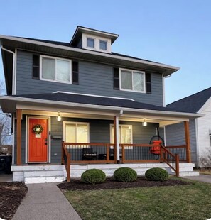 A two-story house with a front porch, a red door, and a small attached garage. - Spacious 5BR  Indy Stay 5 mins to  Downtown -Game Day Deals -HotTub - Firepit (Indianapolis)