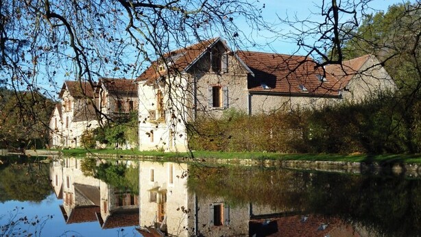 A historic stone building with a reflection in the water, surrounded by trees and a clear sky.