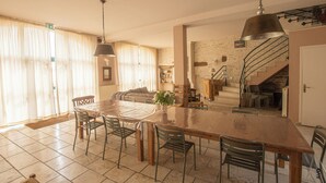 A spacious dining area with a large wooden table and chairs, a staircase leading to an upper level, and a view of the outdoors through large windows.